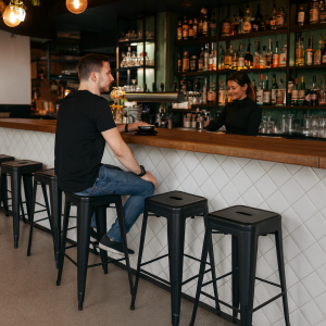 Super-Seat | Toxor Tabouret de bar pour terrasse noir mat | Zwarte metalen barkruk, houten bar en glazen flessen op de achtergrond. | Horecameubilair