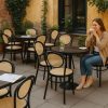 Super-Seat | Une femme est assise seule à une table de café sur une chaise Chopin Noir Thonet (terrasse) avec un cadre en bois, des tables de terrasse en métal, un verre d'eau ; murs jaunes, plantes vertes. | Mobilier pour la restauration