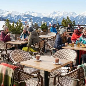 Super-Seat | Chaises beiges boho avec accoudoirs, tables et tabourets en bois sur une terrasse enneigée, personnes en vêtements d'hiver. | Mobilier de restauration