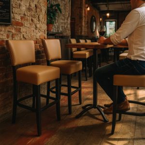 Super-Seat | Une personne déguste un verre de vin rouge à une table haute dans un restaurant rustique | Barkrukken taupe stof en metaal, houten tafels, bakstenen muren, warme verlichting. | Horecameubilair