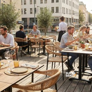 Super-Seat | HPL Hospitality Table Tops Castle Oak 80x80cm Tables en bois de couleur chêne avec châssis et chaises en acier, entourées de plantes et de lumières. | Mobilier de restauration