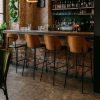 Super-Seat | Un bar avec un sol en pierre, un mur en briques et quatre tabourets de bar design new-yorkais en bois et cuir noir. | Quatre tabourets de bar en cuir noir avec des pieds en bois ; bar avec sol et mur intérieur en pierre. | Mobilier de restauration