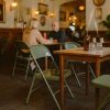 Super-Seat | Deux personnes sont assises l'une en face de l'autre à une table en bois dans un café à la lumière tamisée | Houten tafels, groene metalen vintage klapstoelen, bruine glazen fles decor |. | Horecameubilair